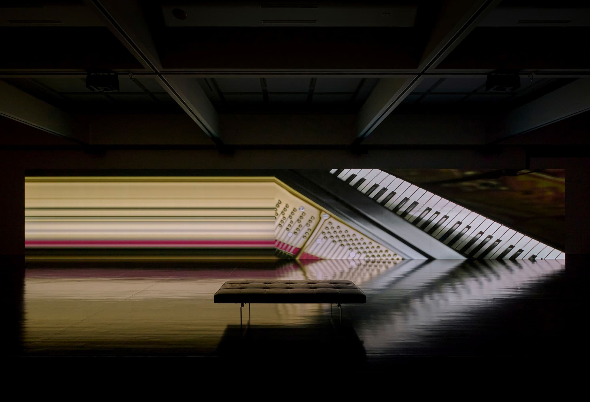 A dark gallery with a projected close-up of piano keys and a bench centered before Ryuichi Sakamoto and Shiro Takatani's async-immersion installation