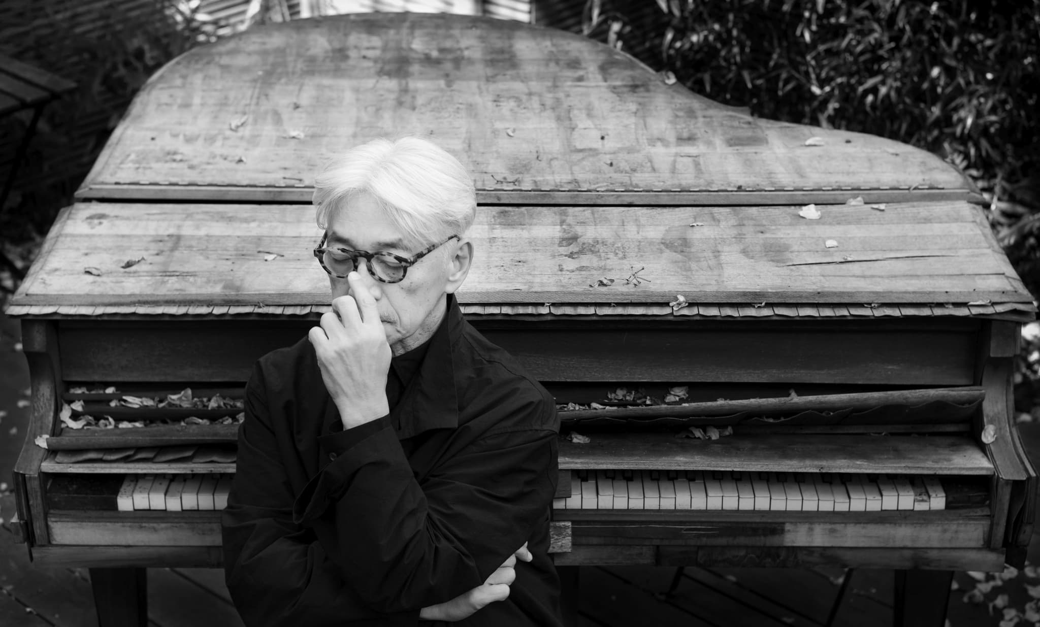 Black-and-white portrait of Ryuichi Sakamoto seated in thought before a weathered outdoor piano, photographed by Neo Sora